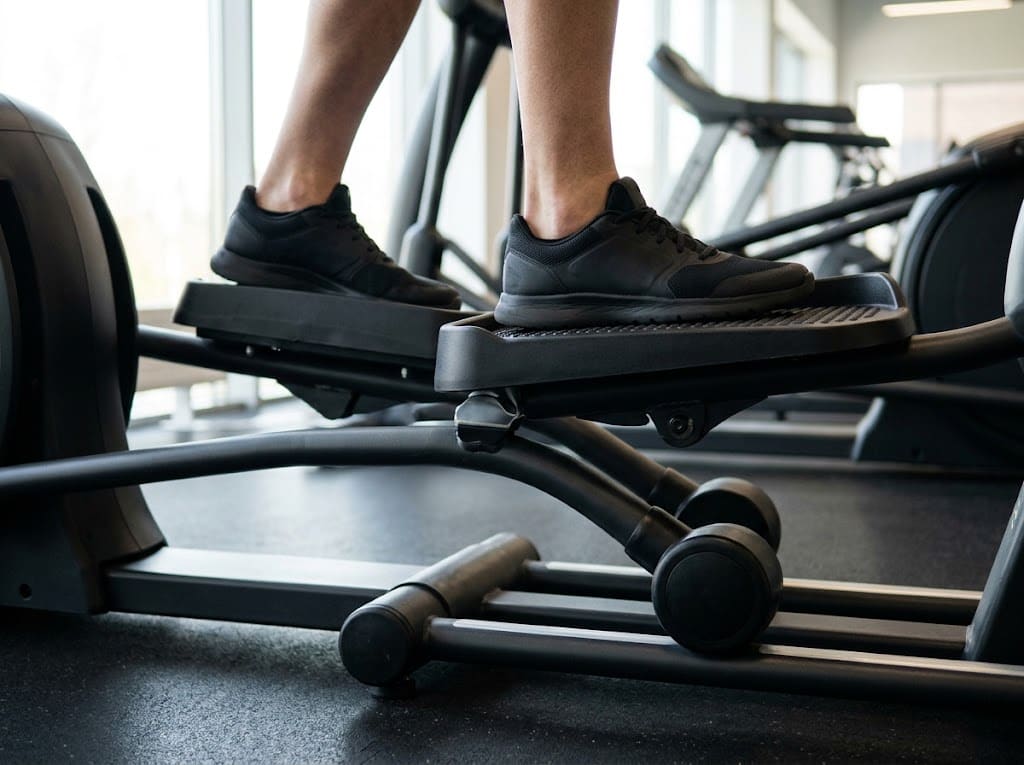 Zbliżenie z boku na stopy w sportowych butach stojące płasko na dużych, antypoślizgowych pedałach orbitreka, pięty dociśnięte, widoczny wąski…