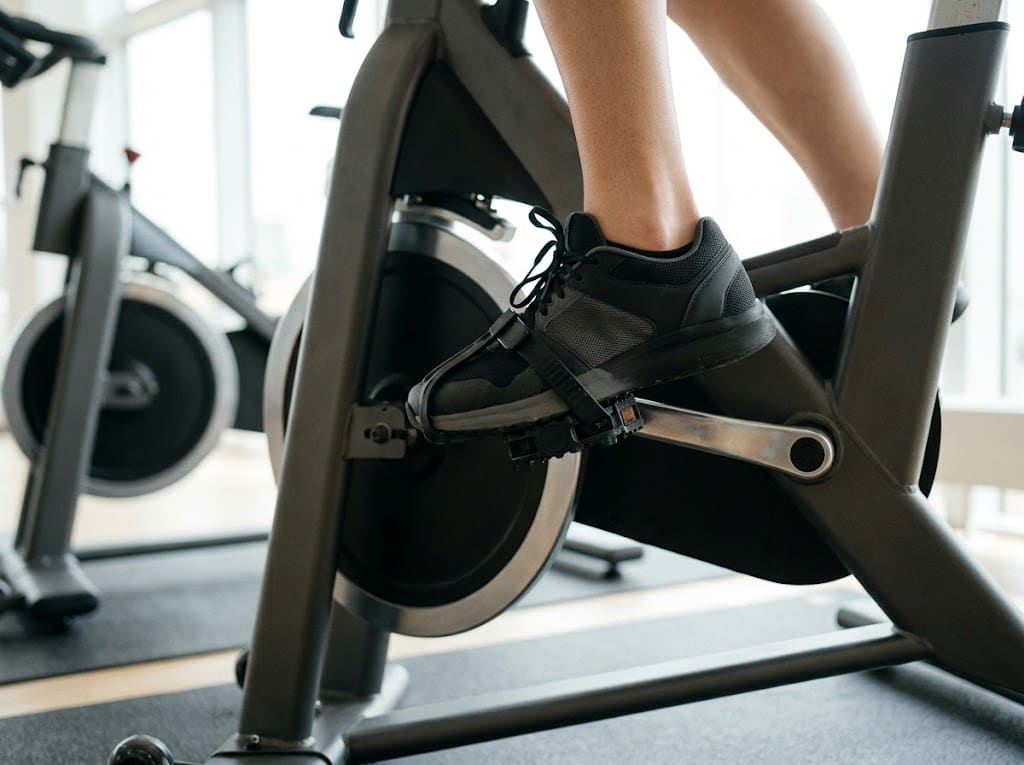 Zbliżenie z boku na stopę w sportowym bucie na pedale rowerka stacjonarnego z dociągniętym paskiem, śródstopie nad osią pedału; w tle rozmyta rama…